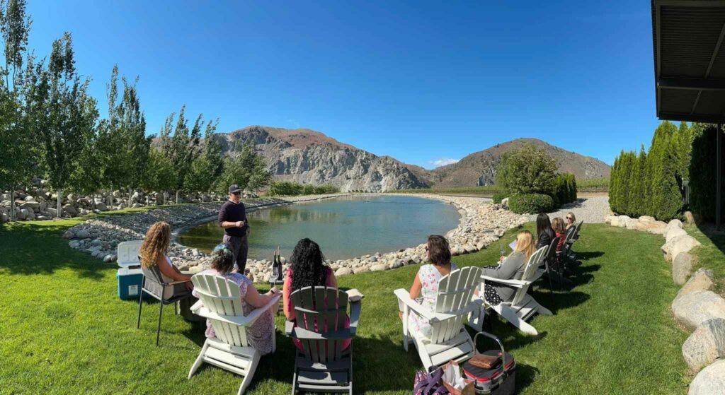 A group of people sit on lawn chairs in front of a small lake with rocky edges, surrounded by trees and mountains under a clear blue sky.