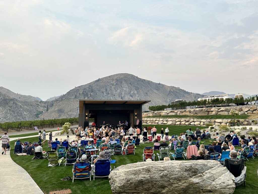 A crowd in foldable chairs sits on a grassy lawn facing an outdoor stage with mountains in the background.