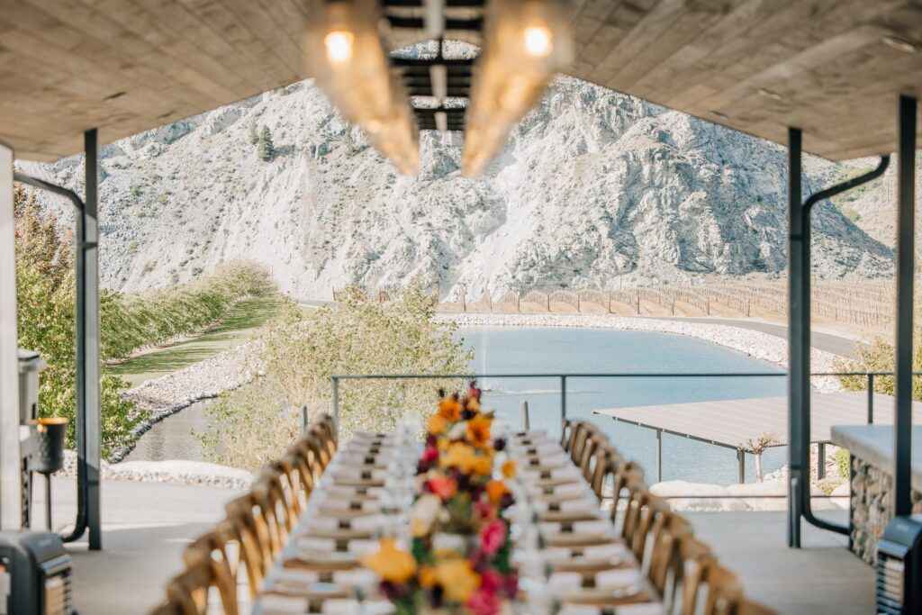 A long wooden table adorned with colorful floral arrangements is set beneath a rustic, open-air pavilion, overlooking a serene pond and rocky landscape.