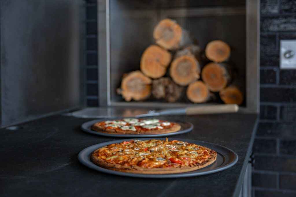 Two pizzas on metal trays sit on a countertop near a wood-fired oven with stacked logs in the background.