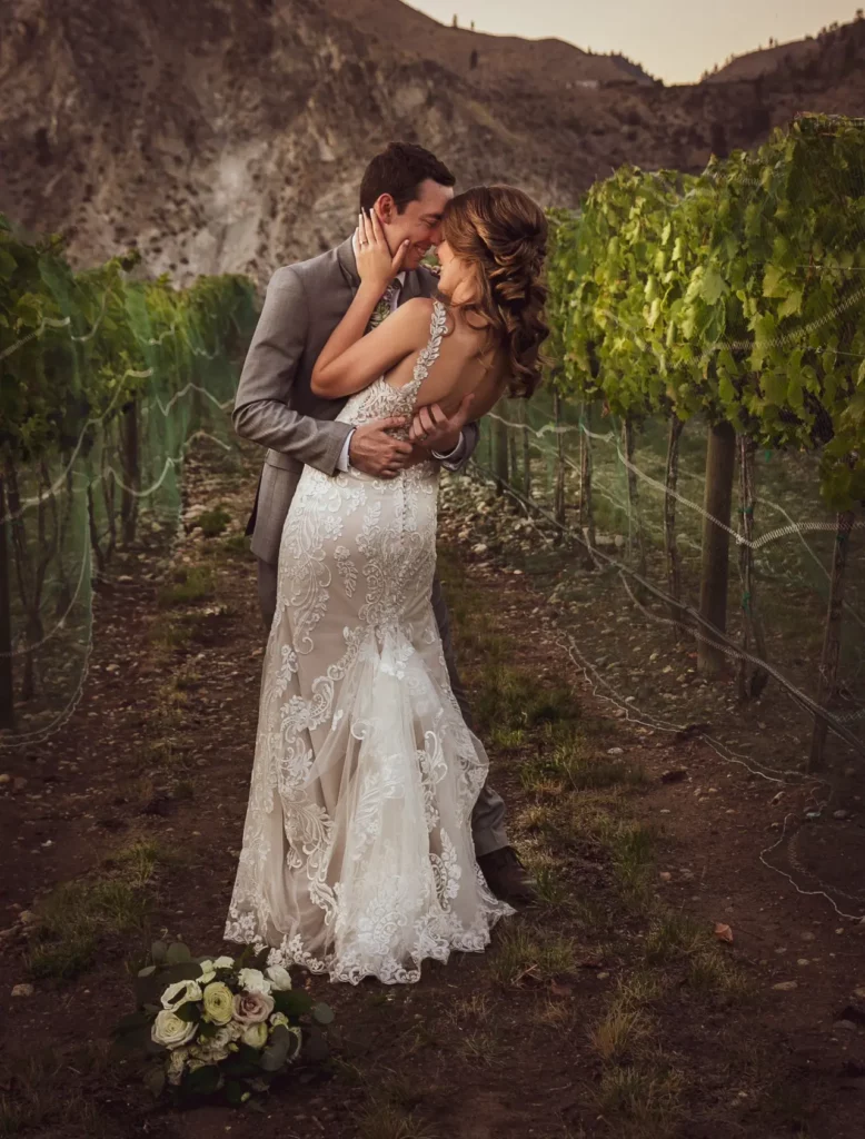 A couple, dressed in wedding attire, stands close together in a vineyard. The bride holds a bouquet and the groom wears a suit. The background features mountainous terrain.