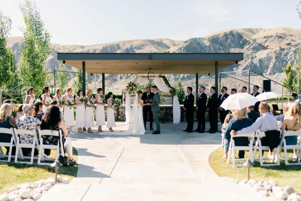 Outdoor wedding ceremony with two people exchanging vows under a canopy, accompanied by bridesmaids and groomsmen, and guests seated on either side in a scenic mountain setting.