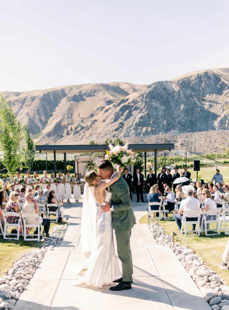 A bride and groom share a kiss at an outdoor wedding ceremony in front of guests, with mountains in the background.