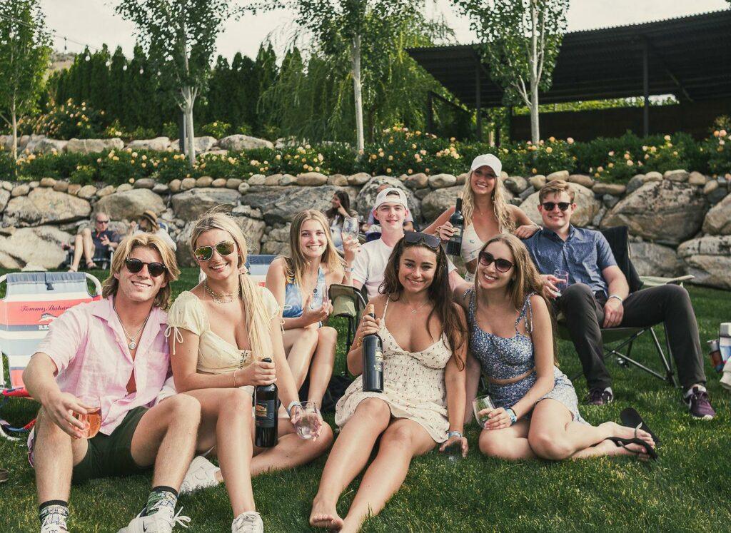 A group of eight young adults sit on grass and enjoy drinks together in an outdoor setting with trees and rocks in the background.