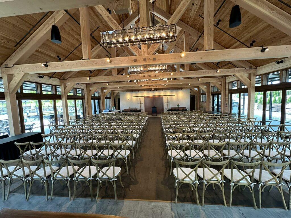 A spacious event hall with exposed wooden beams, rows of white cushioned chairs, two long tables at the front, large windows, and chandeliers.