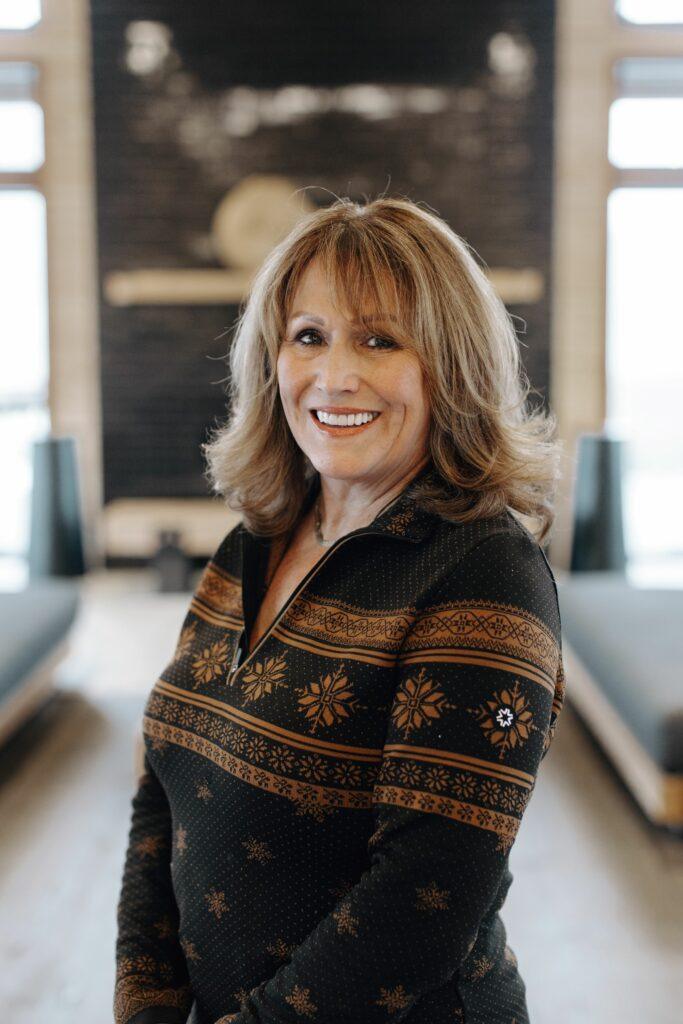 A woman with shoulder-length hair and a patterned sweater smiles while standing in front of a blurred indoor background.