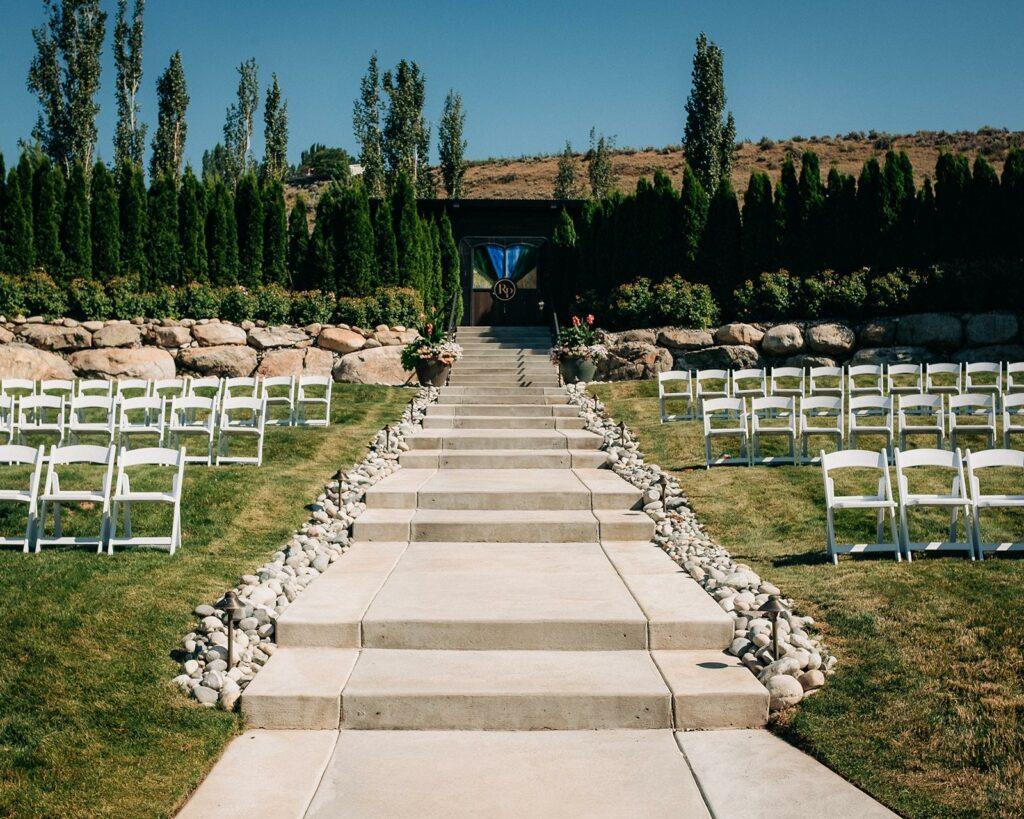 An outdoor wedding venue with rows of white chairs on either side of a stone-paved aisle leading to an altar, surrounded by greenery and tall trees.