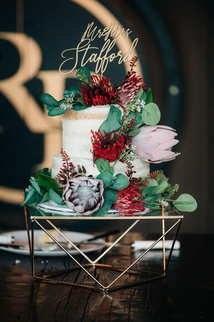 A two-tiered wedding cake adorned with various flowers and greenery, topped with a "Mr. & Mrs. Stafford" decoration. The cake sits on a geometric stand.