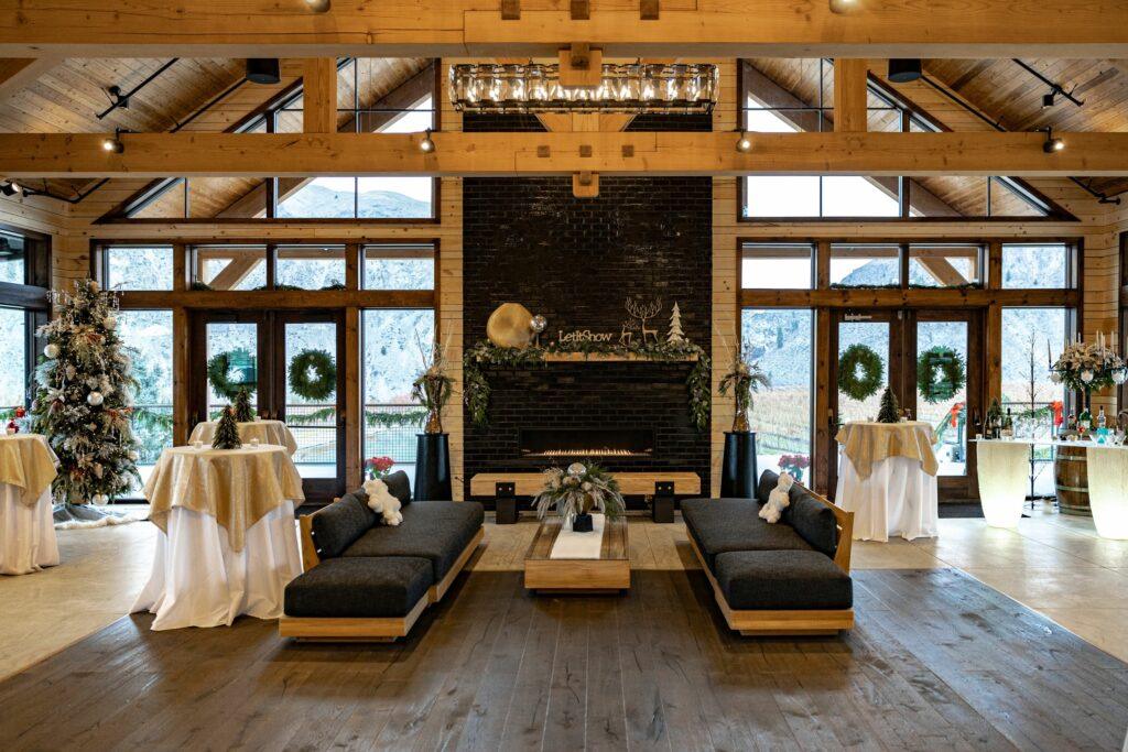 A modern wooden interior decorated for the holidays, featuring a fireplace, large windows, two black sofas, and multiple round tables adorned with white cloths and festive greenery.