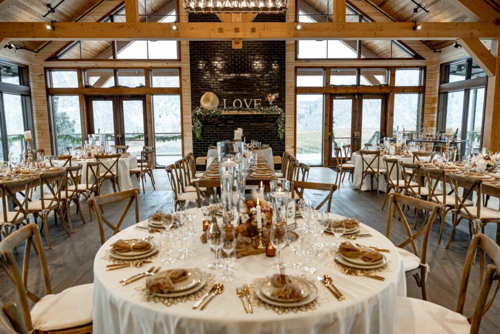 A decorated indoor wedding reception area with round tables set with plates, glasses, and cutlery. The word "LOVE" is displayed on a dark wall in the background.