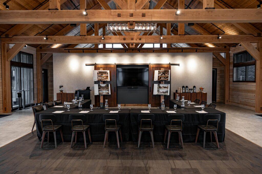 A conference room with a large wooden table arranged in a U-shape, surrounded by chairs. The room has wooden beams, a television screen on the wall, and various decorations and drink stations.