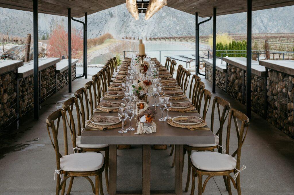 Outdoor dining table set with plates, glasses, and floral centerpieces. Surrounded by wooden chairs, the table is arranged under a covered area with a mountain view in the background.