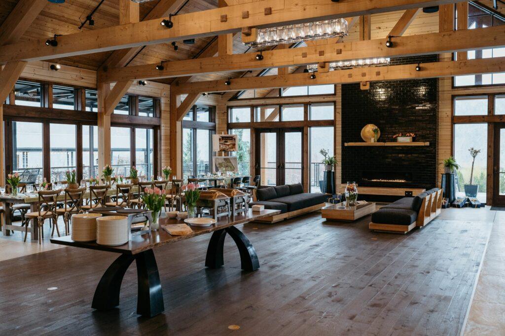 A spacious, wooden interior with large windows, long tables set for a meal, a black fireplace, and seating areas.