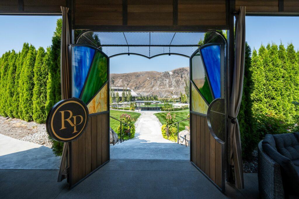 A landscaped garden and mountains are visible through an open, decorative gate bearing the initials "RD." Trees and manicured lawns line the pathway leading to a distant structure.