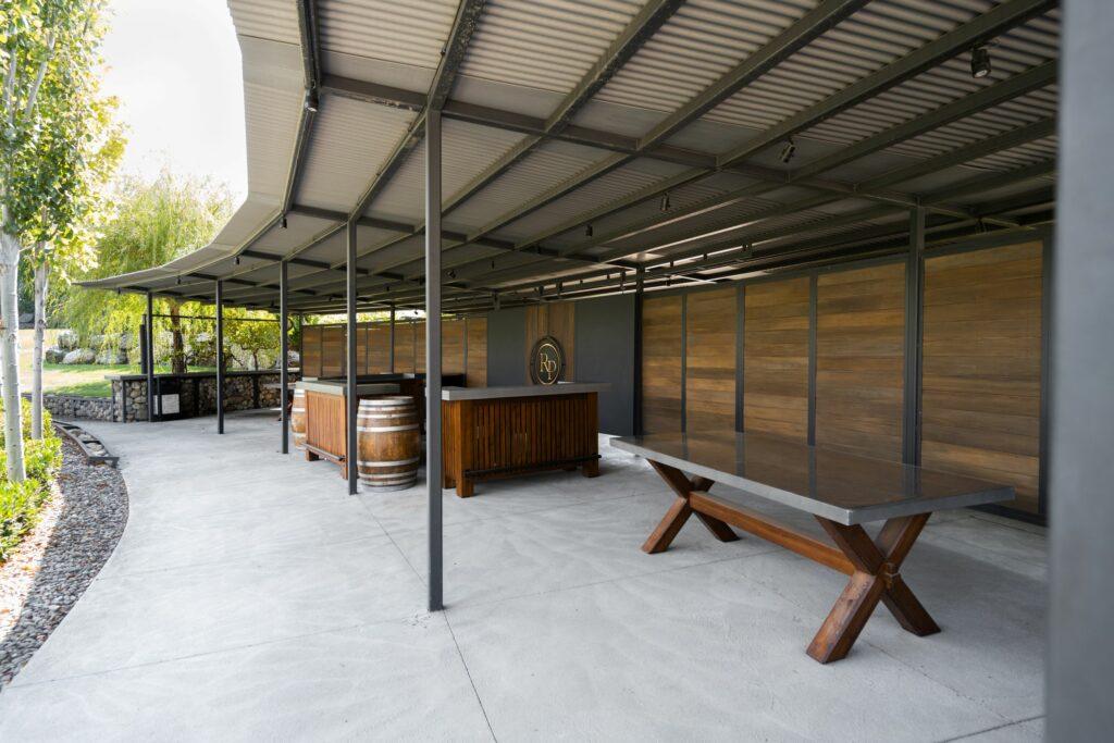 Outdoor pavilion with a covered roof, wooden bar counter, wine barrels, and wooden table on a concrete floor, surrounded by trees and greenery.