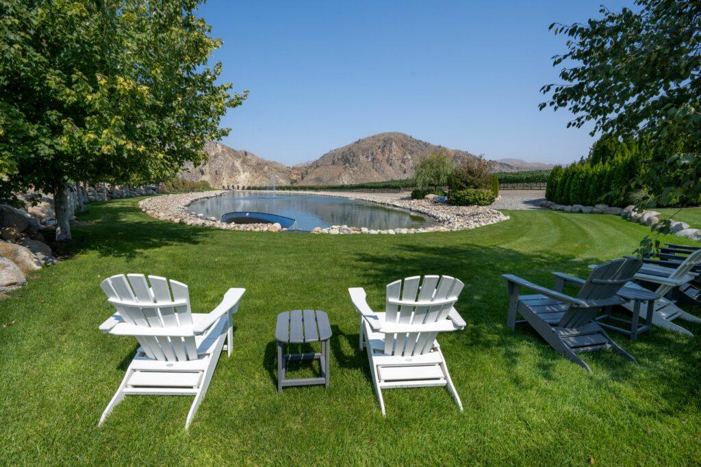 Two white Adirondack chairs and a small table are set on a grassy lawn, facing a serene pond surrounded by rocks, with mountains in the background under a clear blue sky.