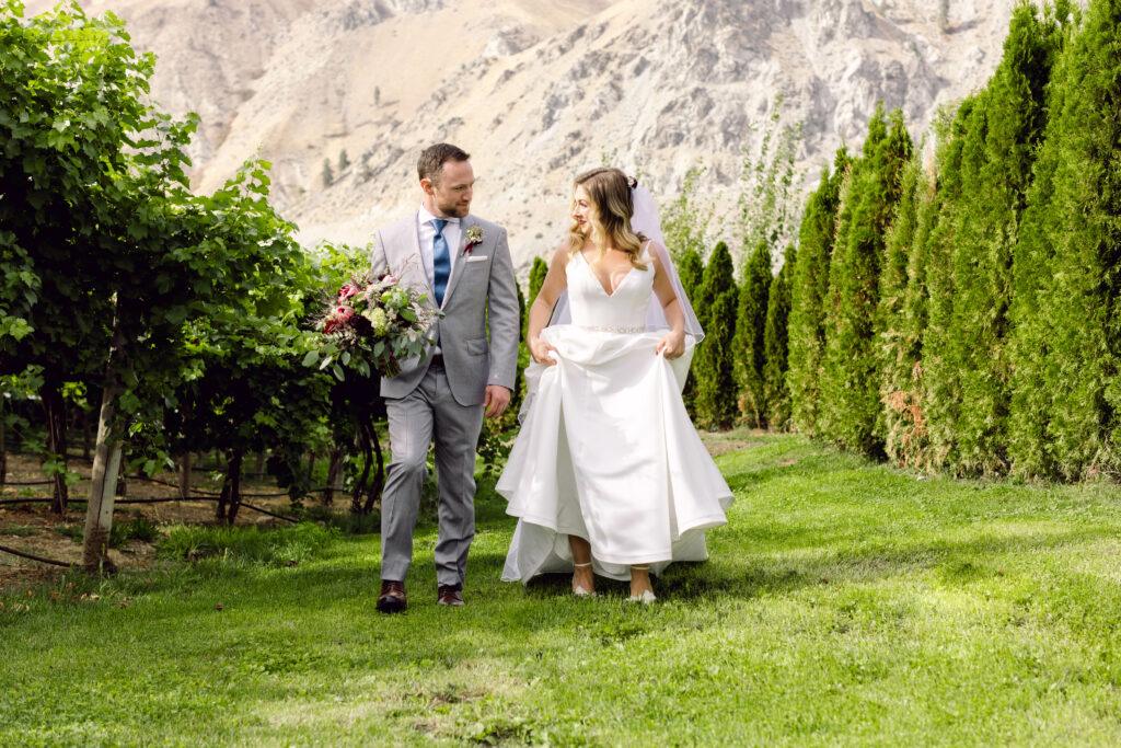 A bride in a white dress and a groom in a gray suit walk hand-in-hand through a vineyard lined with green hedges and trees, with mountainous terrain in the background.