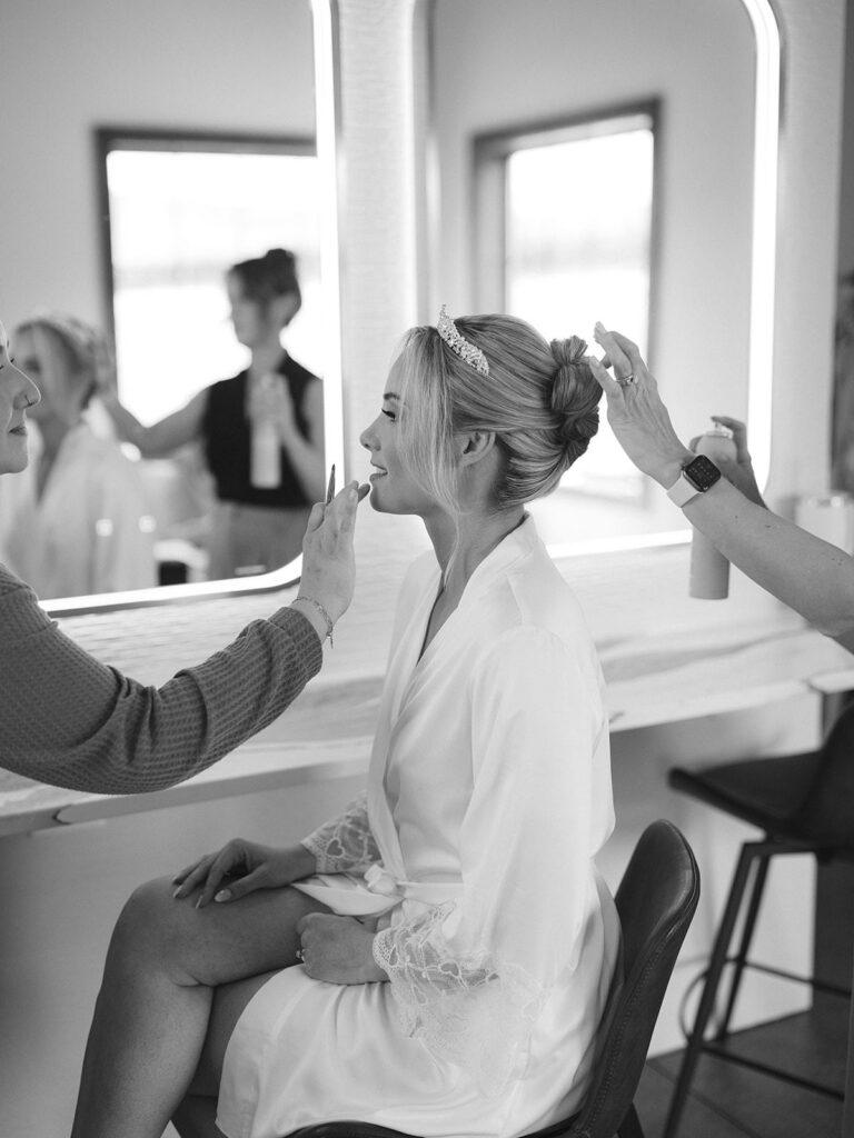 A woman in a robe and tiara is seated, having her makeup applied and hair styled by two people in a salon.