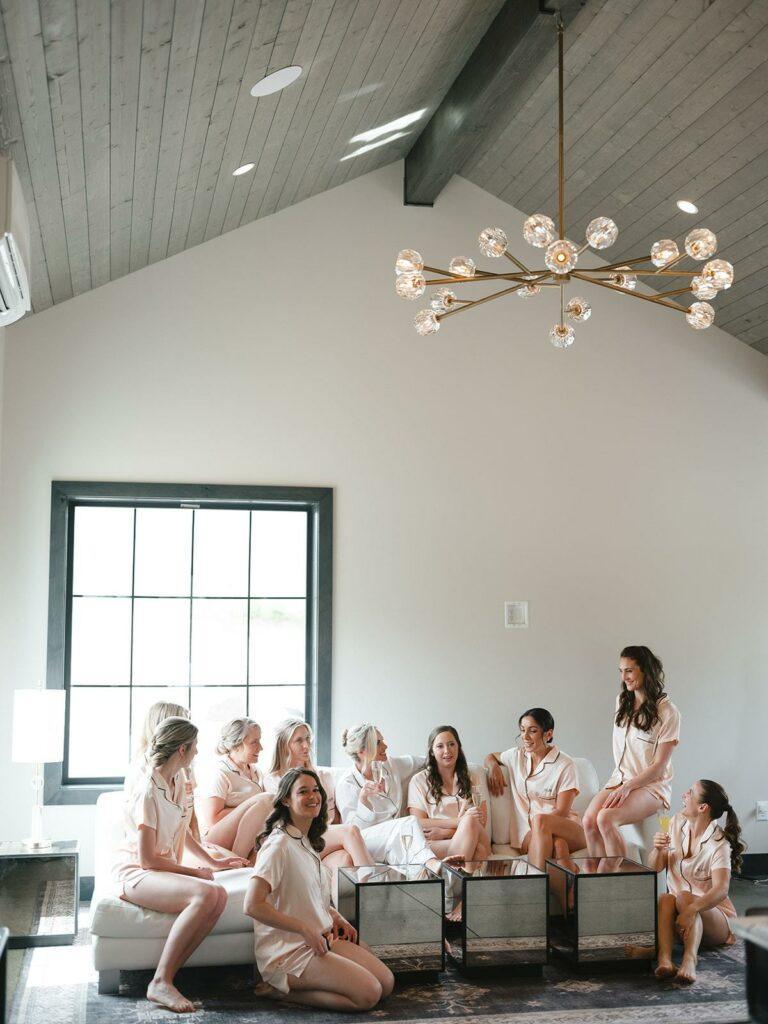 A group of ten women wearing matching light-colored outfits sit and stand around a couch in a modern, well-lit room with a high ceiling and a large window.