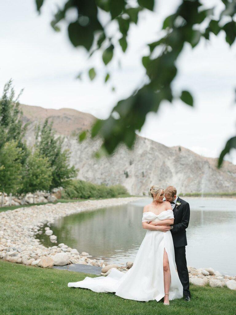 A couple stands by a lake with mountains in the background. The person in a white dress is embraced by the person in a black suit. Trees and rocks are visible around the lake.