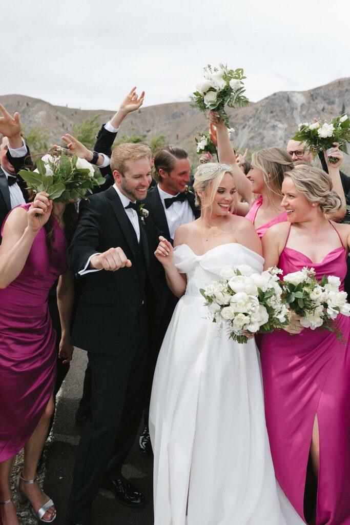 A newlywed couple smiles and celebrates with their wedding party. The bride is in a white dress, the groom in a black suit, and the bridesmaids wear magenta dresses and hold bouquets.