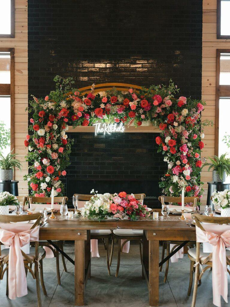 Elegant wooden dining table set for an event, adorned with floral centerpieces. A large floral arch with a neon sign reading "The Kitchen" is in the background against a dark brick wall.