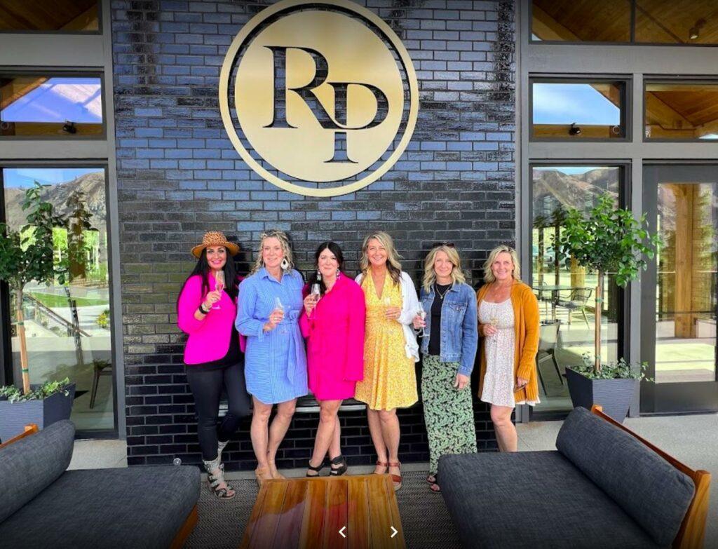 Six women standing in front of a black brick wall with a large circular "RD" logo, holding wine glasses. They are dressed in colorful clothing and smiling at the camera.