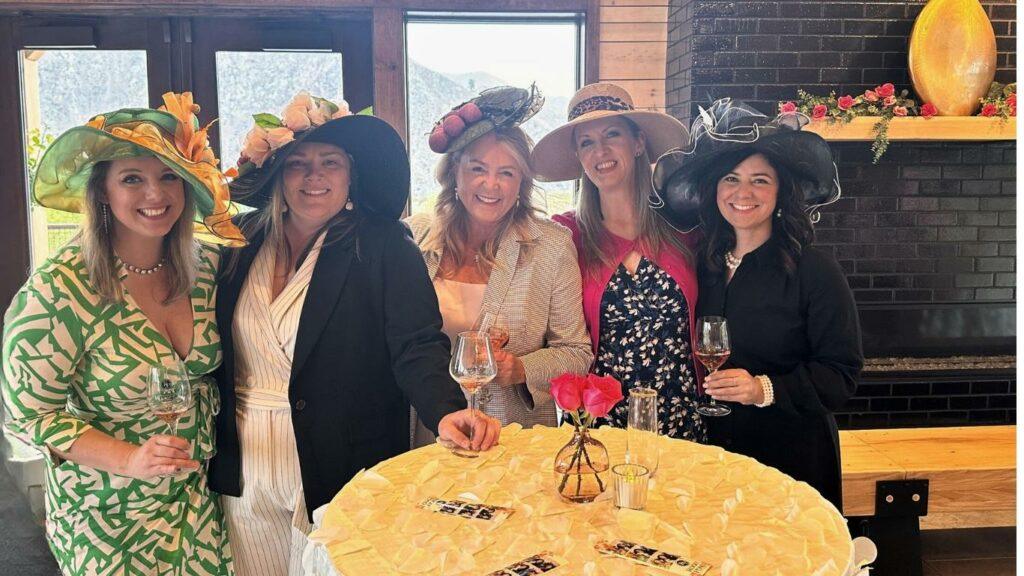 Five women are standing around a table, each holding a glass, and wearing decorative hats. They are smiling and posing for the photo. The table has a small vase with a flower. The background includes a fireplace.