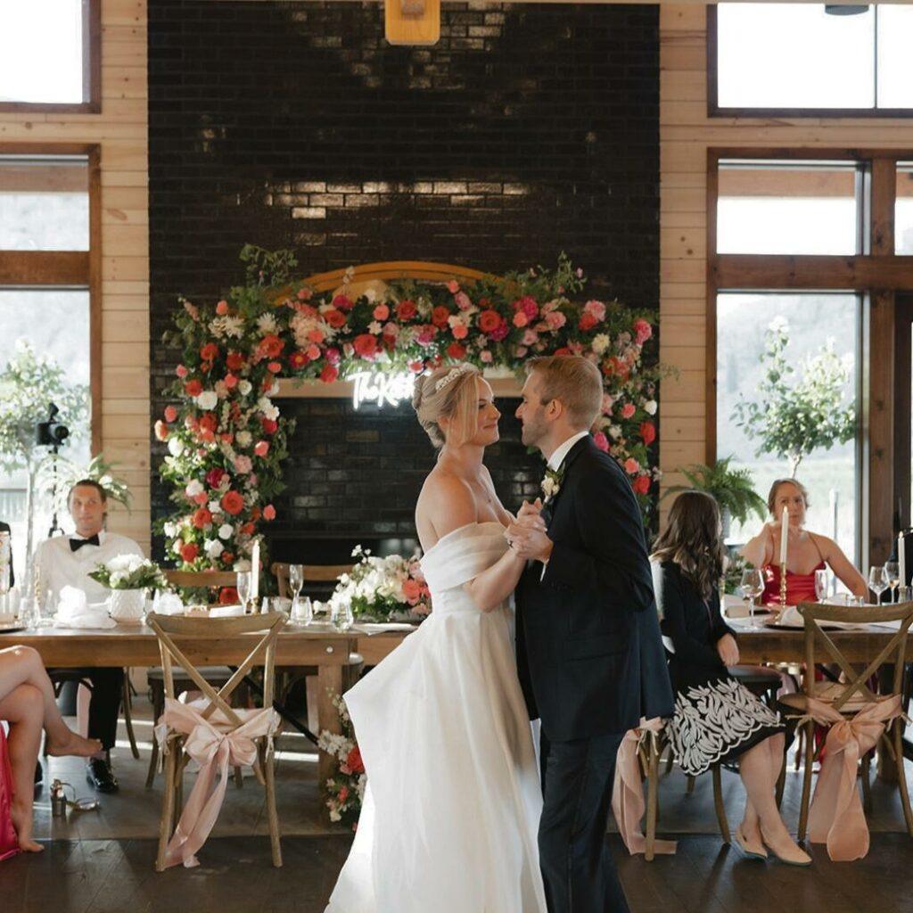 A bride and groom share a first dance in front of a floral arch, with wedding guests seated at a long wooden table in the background.