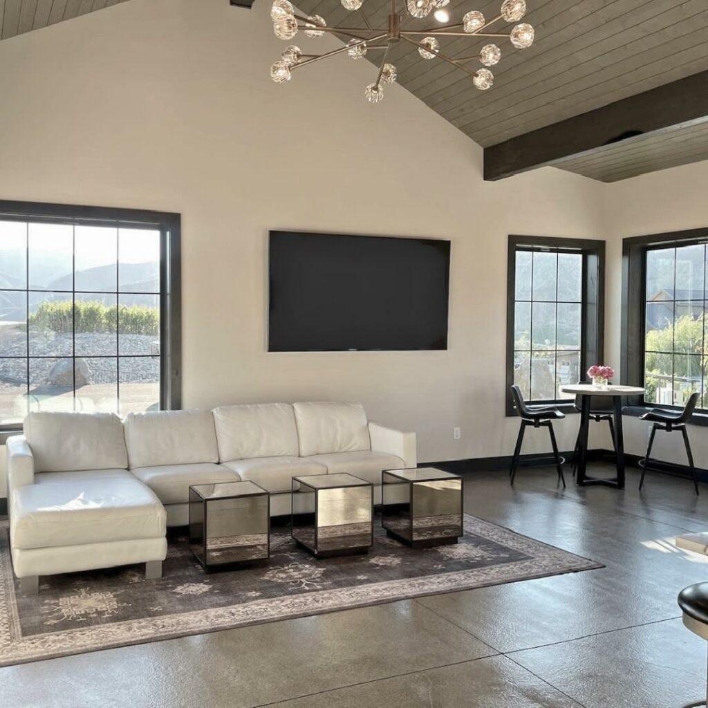 A modern living room with a white sectional sofa, three glass cube coffee tables, a wall-mounted TV, and large windows offering views of hills. A small round dining table with two chairs is in the corner.