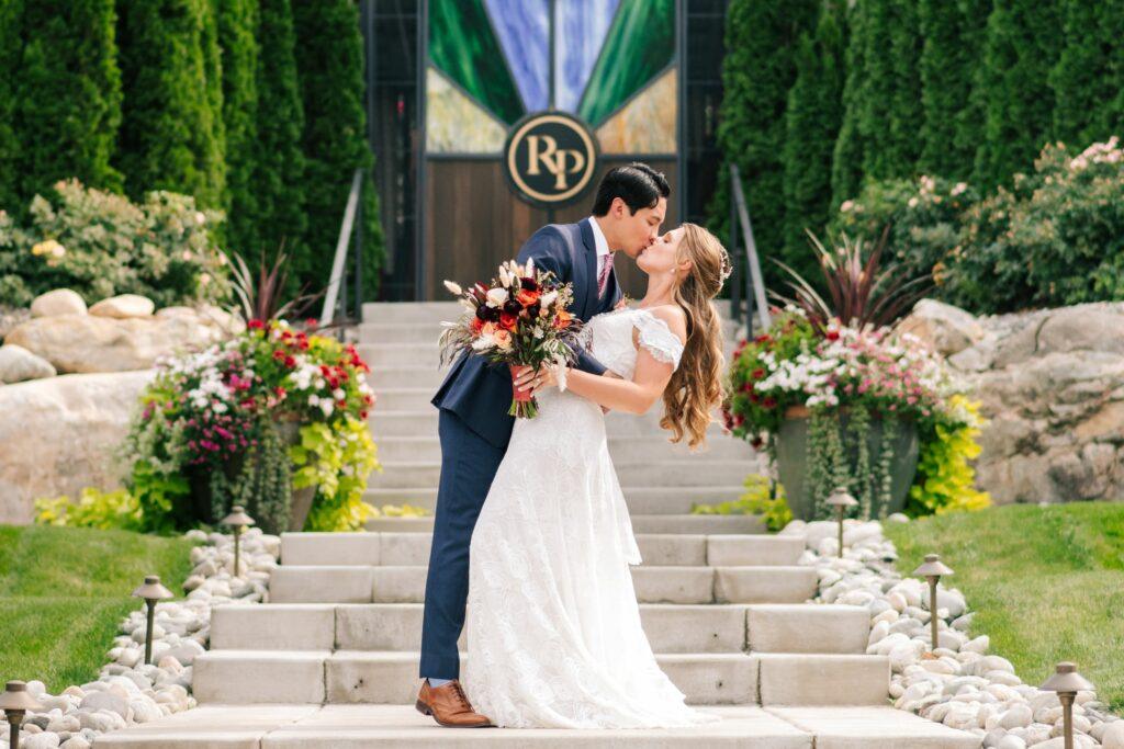 A couple in wedding attire passionately kiss on an outdoor staircase, surrounded by vibrant greenery and flowers, with a decorative door and an 'RP' emblem in the background.