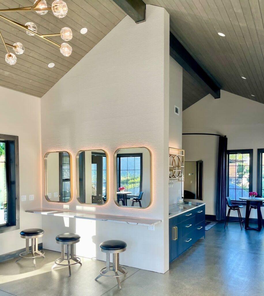 Modern open-concept room with vaulted ceiling, four illuminated mirrors above a counter with three stools, a blue cabinet with drawers and a sink, and a dining area visible in the background.