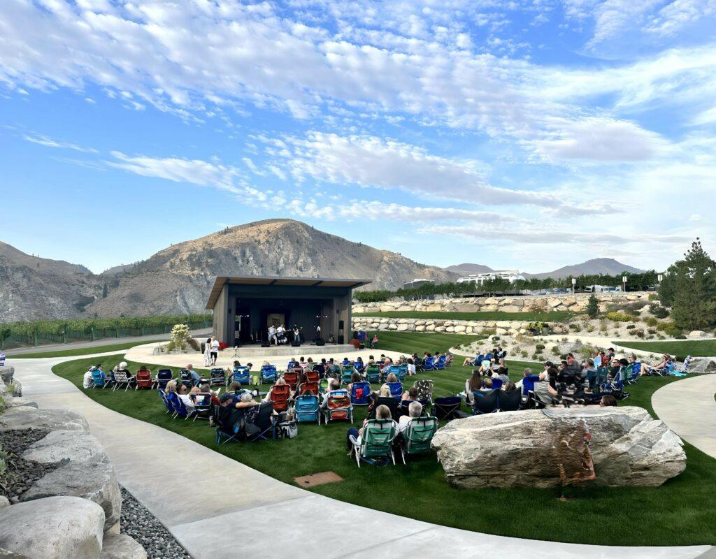 People sitting on lawn chairs and blankets in an outdoor amphitheater enjoying a live performance with mountains in the background under a partly cloudy sky.