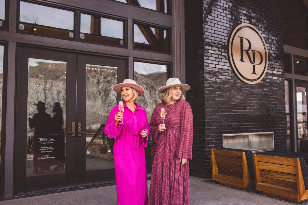 Two women in pink and burgundy dresses, wearing hats, stand outside a building with a large "RD" sign. They are holding champagne glasses and smiling, with a reflection of the mountains in the glass doors.