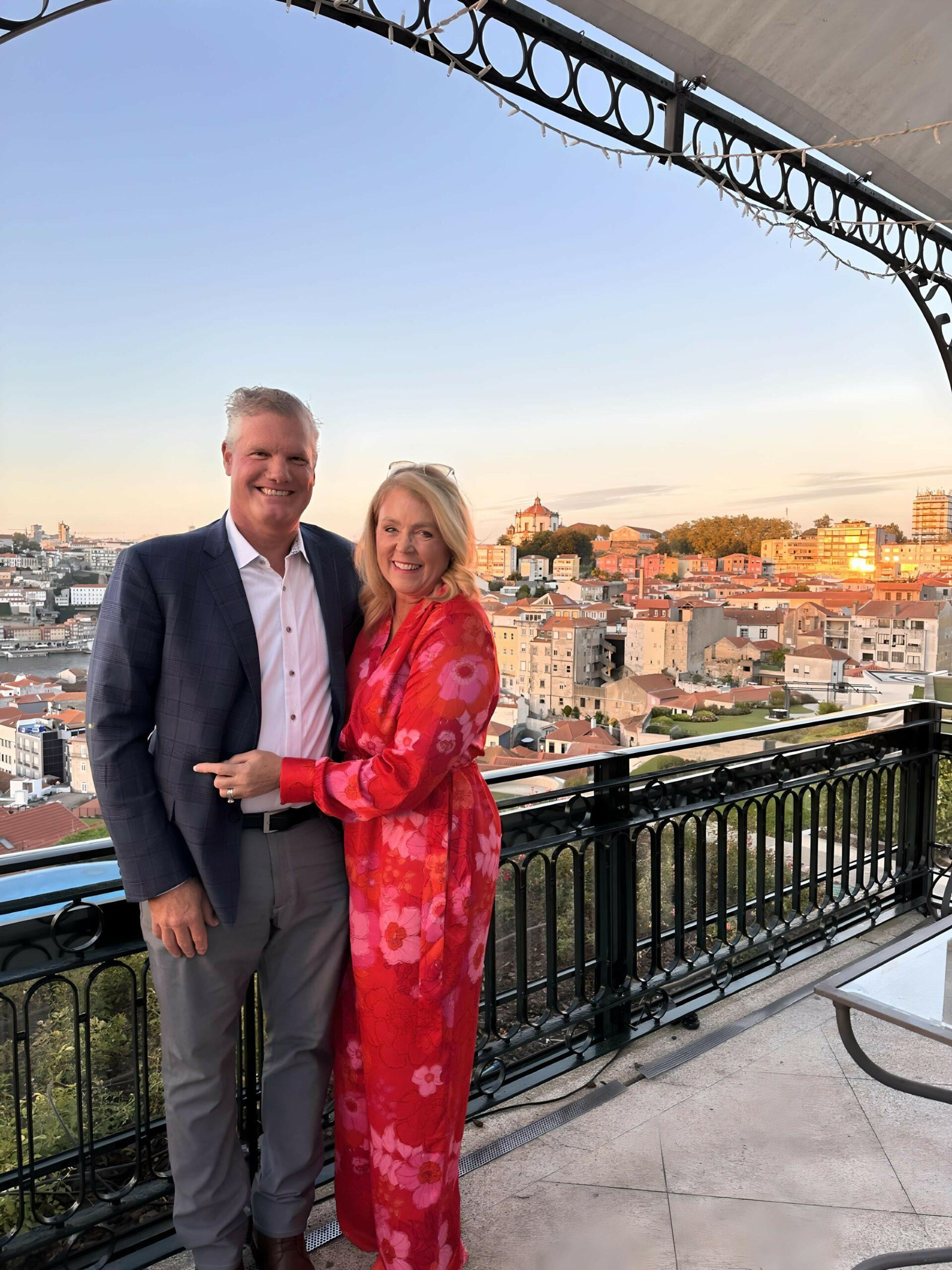 A man and woman pose together on a terrace with a cityscape in the background at sunset. The woman wears a red floral dress, and the man wears a blazer and trousers.