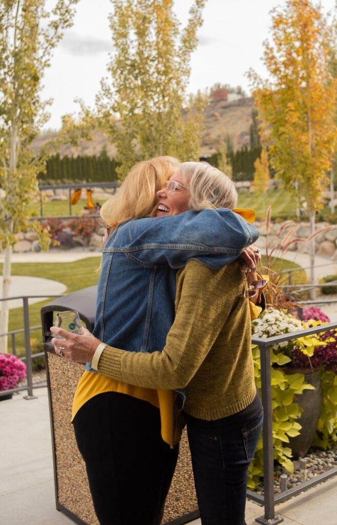 Two women embrace and smile outdoors, surrounded by autumnal trees and plants. One holds a smartphone.