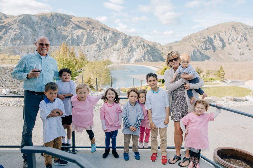 A group photo features two adults, one holding a toddler, and nine children standing in front of a mountainous landscape with a river in between. The children wear pink and gray shirts.