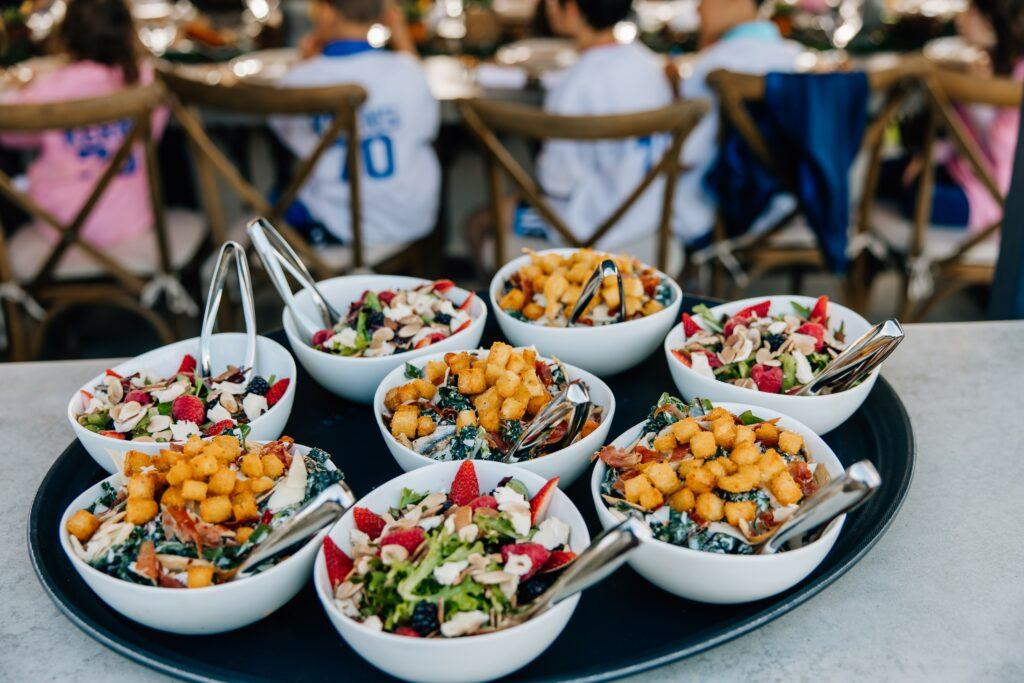 A variety of salads topped with croutons and fruits are served in white bowls on a large tray, with people sitting at tables in the background.