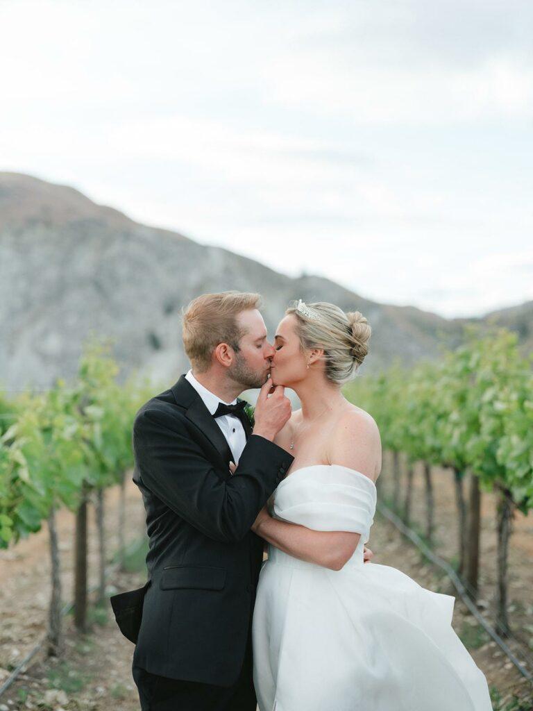 A bride and groom share a kiss in a vineyard, with mountains in the background. The bride is in an off-shoulder white dress, while the groom wears a black tuxedo.